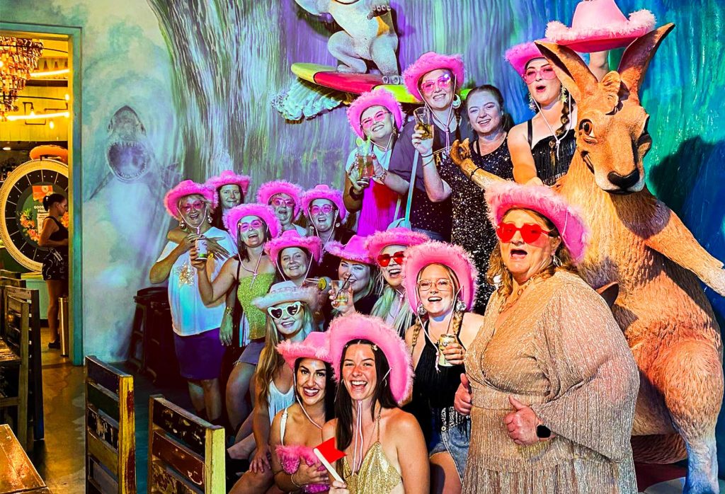 A group of women posing for a photo in front of the surfing kangaroo statue with pink glasses and pink cowboy hats on